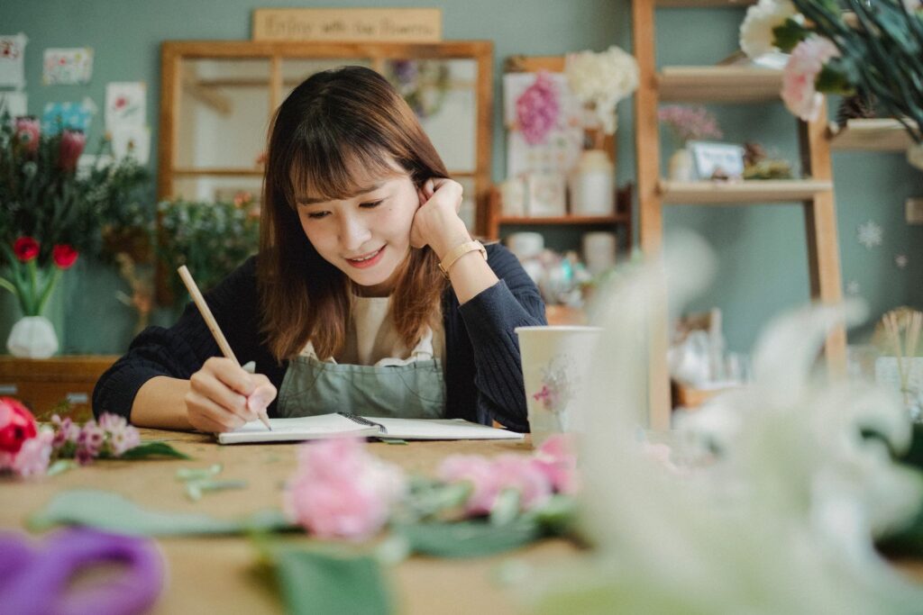 Smiling young female florist in apron writing notes while working in cozy flower shop